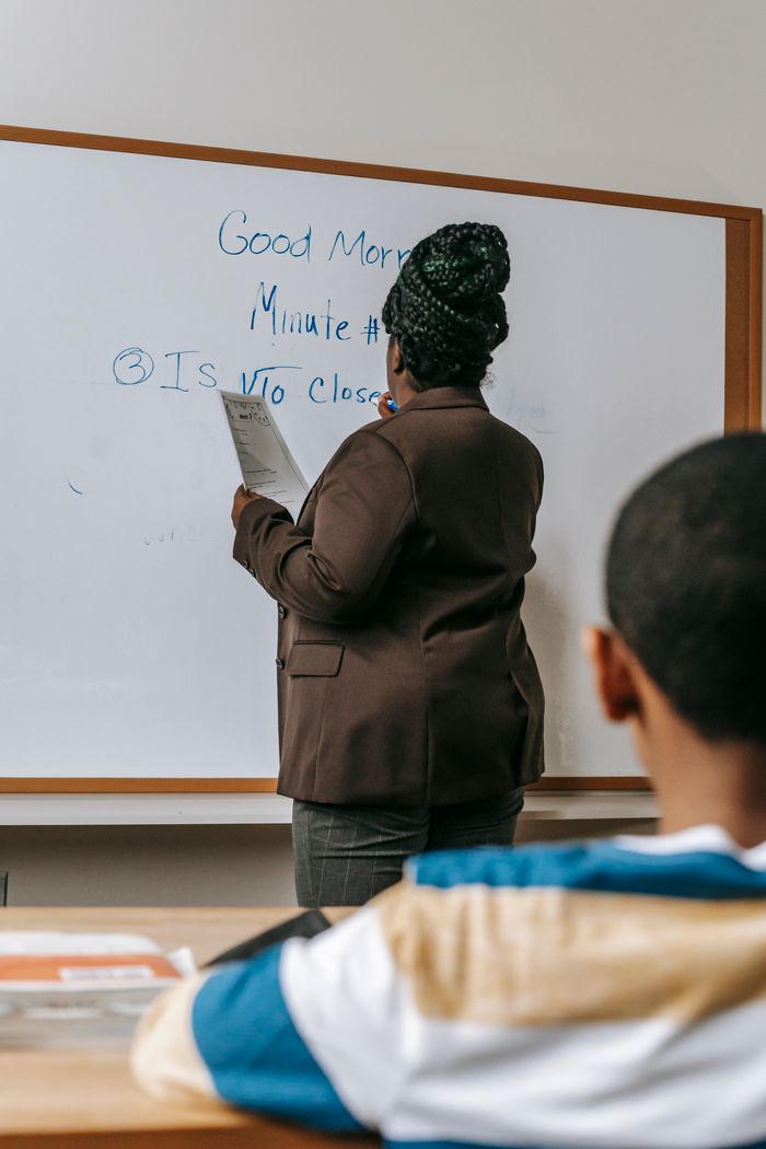 Back view anonymous African American schoolboy sitting at desk in classroom and listening to female teacher taking notes on whiteboard